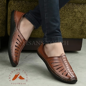 May include: Brown leather sandals with a cut-out design. The sandals have a black sole and are worn by a person with blue jeans. The sandals are labeled "Artisans Touch Footwear".