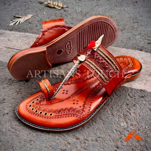 May include: Handmade orange leather sandals with intricate embossed designs. The sandals feature a toe loop, decorative straps, and a textured sole. The image includes the text "ARTISANS TOUCH FOOTWEAR".