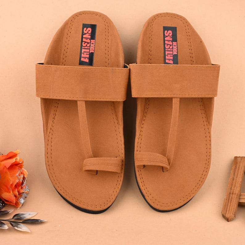Op de afbeelding: Een paar bruine leren sandalen met een teenlus en een verstelbare band. De sandalen hebben de tekst "ARTISANS TOUCH" op de band gedrukt.