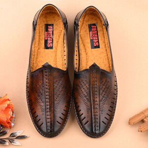K&ouml;nnte beinhalten: Ein Paar braune Slipper aus Leder mit perforiertem Design und dem Schriftzug "Artisans Touch" auf der Innensohle.