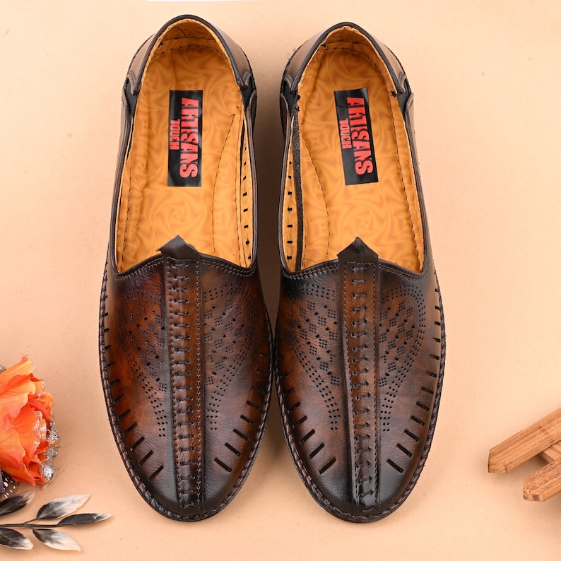 K&ouml;nnte beinhalten: Ein Paar braune Slipper aus Leder mit perforiertem Design und dem Schriftzug "Artisans Touch" auf der Innensohle.
