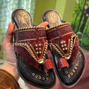 May include: A pair of handcrafted leather sandals in shades of burgundy and black, adorned with intricate gold-colored embellishments. The sandals feature a toe loop and decorative stitching, showcasing a traditional design.