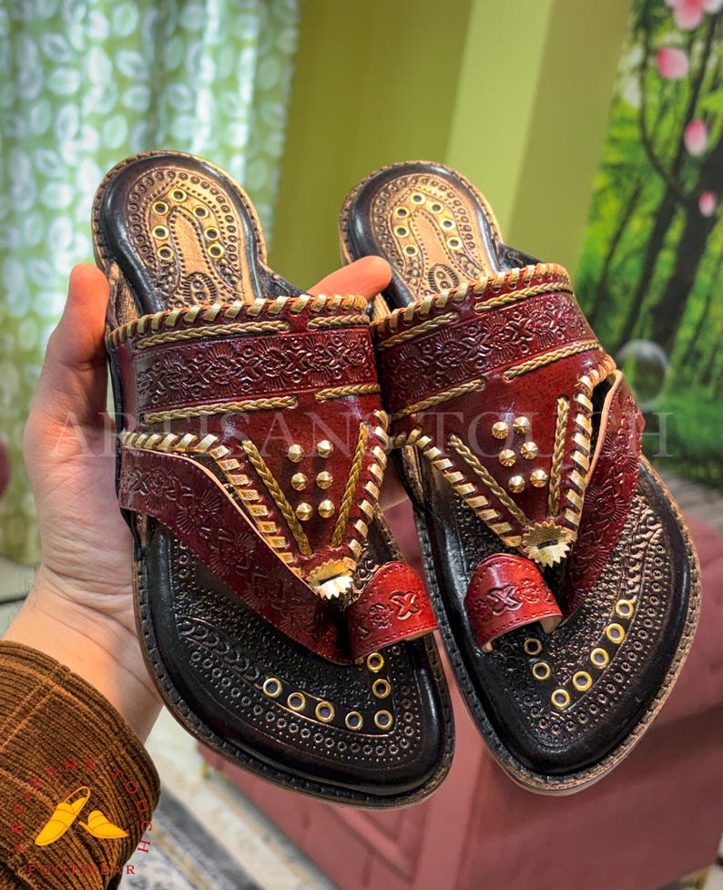 May include: A pair of handcrafted leather sandals in shades of burgundy and black, adorned with intricate gold-colored embellishments. The sandals feature a toe loop and decorative stitching, showcasing a traditional design.