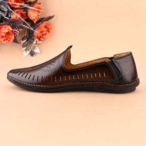 K&ouml;nnte beinhalten: Braune Lederschuhe zum Hineinschl&uuml;pfen mit perforiertem Design und einem schwarzen elastischen Riemen an der Ferse.