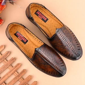K&ouml;nnte beinhalten: Ein Paar braune Slipper aus Leder mit perforiertem Design und dem Schriftzug "Artisans Touch" auf der Innensohle.