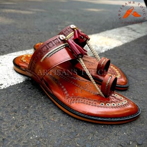 May include: A pair of handcrafted brown leather sandals with intricate embossed designs. The sandals feature a toe loop, decorative tassels, and the text "ARTISANS TOUCH FOOTWEAR". The footwear is displayed on a gray surface.