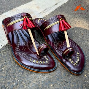 May include: A pair of burgundy leather sandals with intricate embossed patterns and gold accents. The sandals feature a toe loop, decorative tassels, and a gold trim. The soles have an orange border and the text "ARTISANS TOUCH FOOTWEAR".
