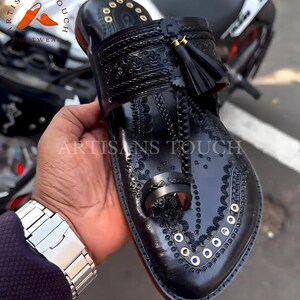 May include: Black leather sandal with intricate embossed floral patterns and gold-colored accents. Features a toe loop, a decorative tassel, and the text "ARTISANS TOUCH FOOTWEAR". The sandal is held in a person's hand.