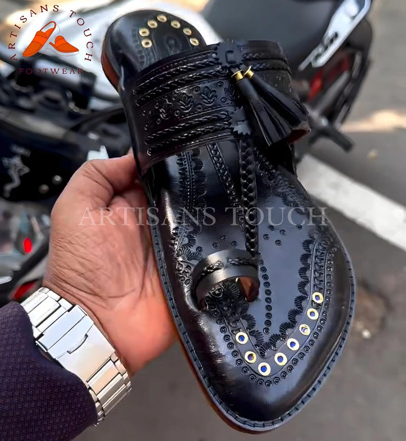 May include: Black leather sandal with intricate embossed floral patterns and gold-colored accents. Features a toe loop, a decorative tassel, and the text "ARTISANS TOUCH FOOTWEAR". The sandal is held in a person's hand.