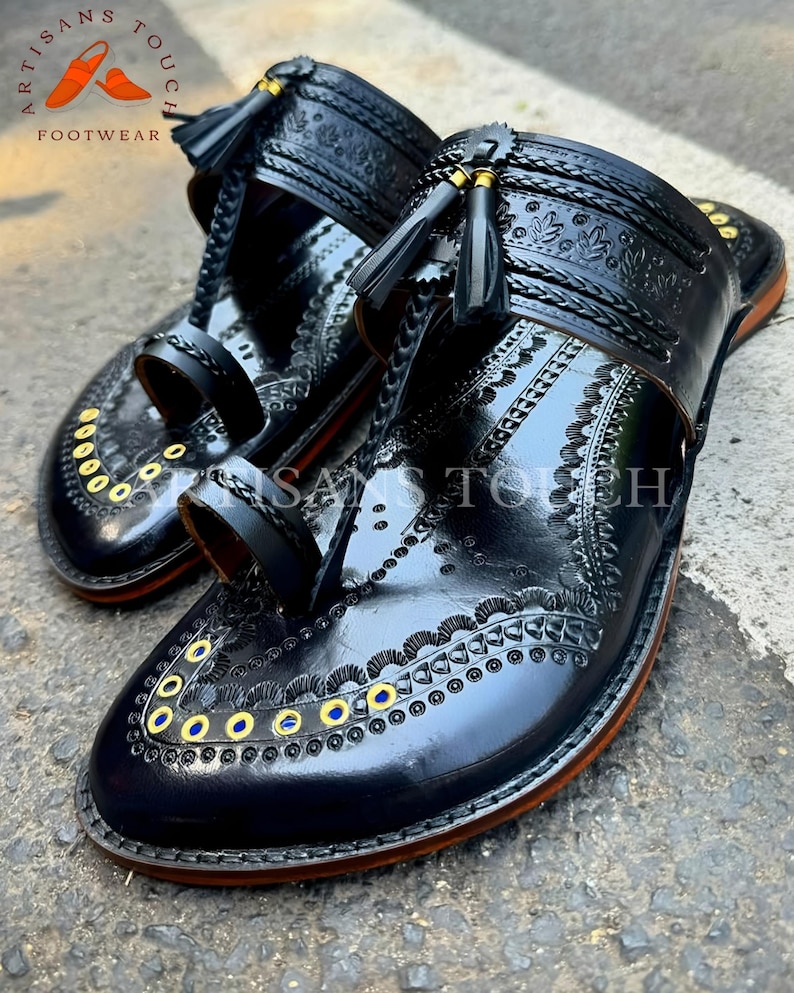 May include: A pair of black leather sandals with intricate embossed designs and decorative tassels. The sandals feature a toe loop and a strap across the top of the foot. Gold and blue accents are visible. The sandals are labeled "ARTISANS TOUCH FOOTWEAR".