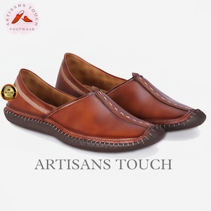 May include: A pair of brown leather slip-on shoes with a dark brown sole and white stitching. The shoes have a rounded toe and are labeled "ARTISANS TOUCH FOOTWEAR".