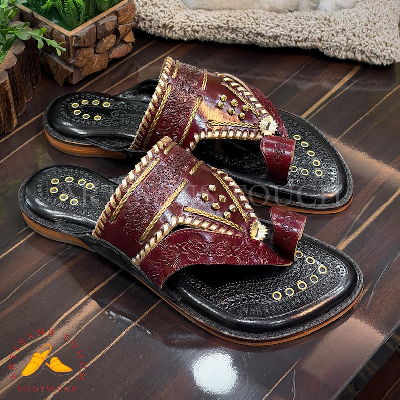 May include: A pair of handcrafted leather sandals. The sandals feature a deep burgundy upper with intricate embossed patterns and gold-colored stitching and studs. The soles are black with decorative circular cutouts. The sandals are displayed on a wooden surface.