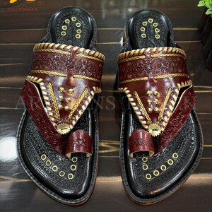 May include: A pair of handcrafted leather sandals. The sandals feature a black base with intricate embossed patterns and a deep burgundy strap. Gold-colored accents and stitching add detail. The sandals are labeled "ARTISANS TOUCH FOOTWEAR".
