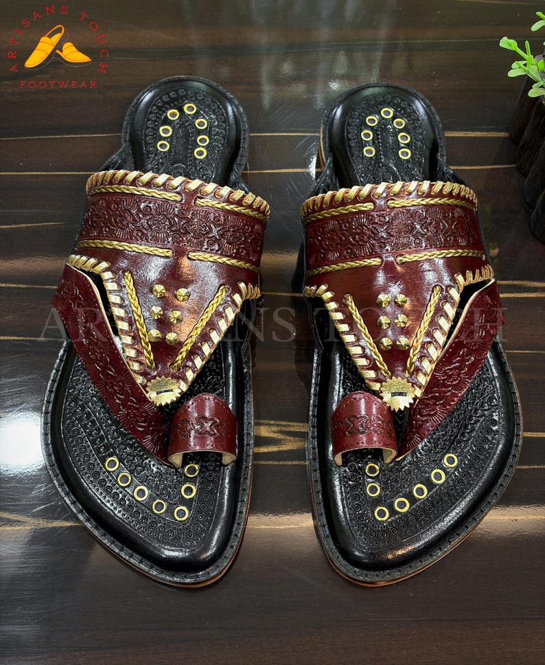 May include: A pair of handcrafted leather sandals. The sandals feature a black base with intricate embossed patterns and a deep burgundy strap. Gold-colored accents and stitching add detail. The sandals are labeled "ARTISANS TOUCH FOOTWEAR".