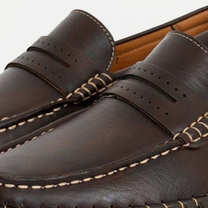 May include: A pair of brown leather loafers with white stitching around the edges. The loafers have a perforated design on the top and a moc toe.