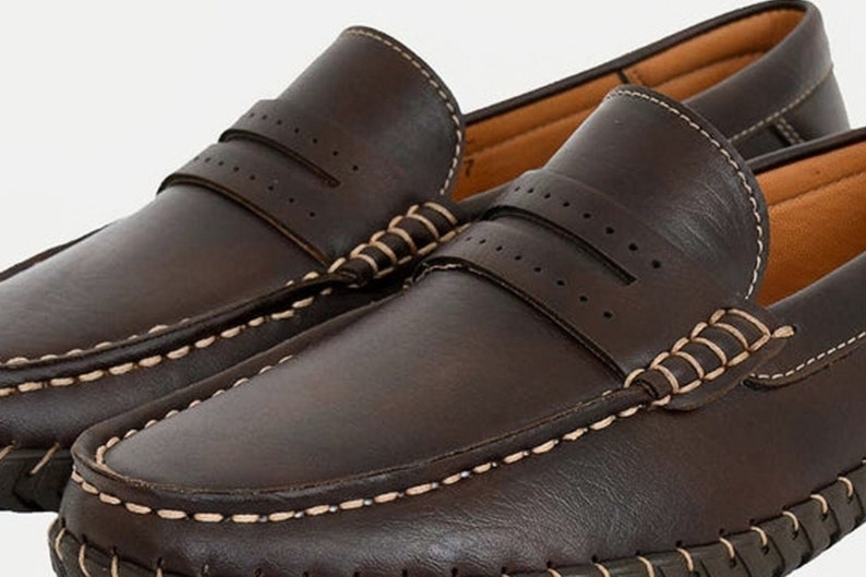 May include: A pair of brown leather loafers with white stitching around the edges. The loafers have a perforated design on the top and a moc toe.