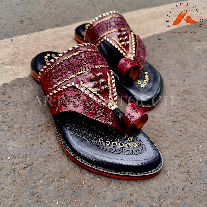 May include: A pair of handcrafted leather sandals. The sandals feature a deep red upper with intricate floral and geometric patterns, gold stitching, and decorative studs. The soles are black with a red trim, and the sandals have a toe loop design.