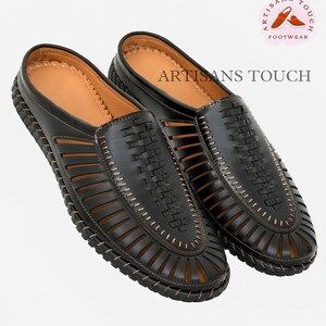 May include: A pair of black leather sandals with a woven design and a brown stitching detail. The sandals have an open toe and a closed heel. The sandals are made by Artisans Touch Footwear.