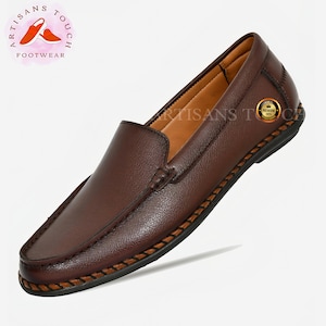 May include: Brown leather slip-on loafers with a stitched edge detail. The shoes have a brown leather lining and a black rubber sole. The brand name "Artisans Touch Footwear" is visible on the image.
