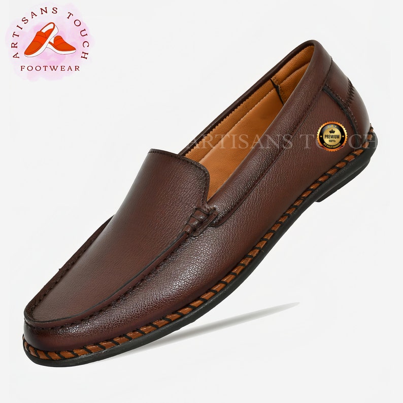 May include: Brown leather slip-on loafers with a stitched edge detail. The shoes have a brown leather lining and a black rubber sole. The brand name "Artisans Touch Footwear" is visible on the image.