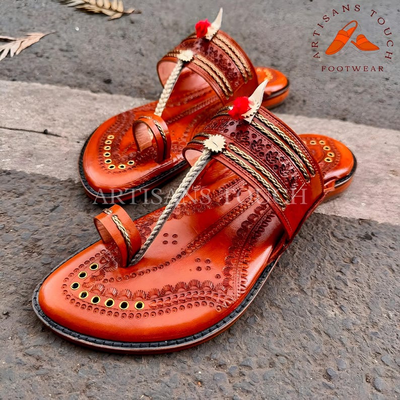 May include: A pair of handcrafted, brown leather sandals with intricate embossed designs. The sandals feature a toe loop, decorative straps, and a rope-like thong. Accented with gold trim and small red pom-poms. The text "ARTISANS TOUCH FOOTWEAR" is visible.