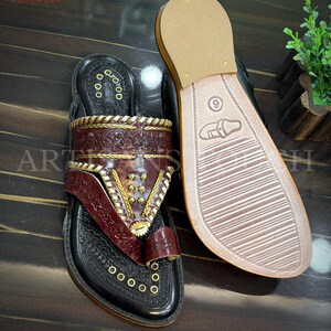 May include: Black leather sandals with intricate burgundy and gold detailing. The sandals feature a toe loop and decorative stitching. The sole of one sandal is visible, displaying a size 9 marking. The sandals are displayed on a dark wooden surface.
