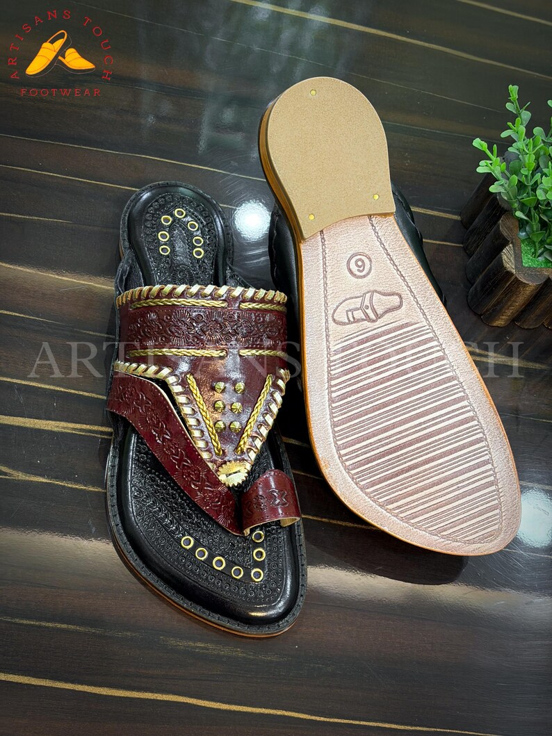 May include: Black leather sandals with intricate burgundy and gold detailing. The sandals feature a toe loop and decorative stitching. The sole of one sandal is visible, displaying a size 9 marking. The sandals are displayed on a dark wooden surface.