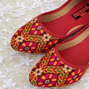May include: Pair of red flat shoes with a colorful floral pattern. The shoes have a pointed toe and are made of fabric. The brand name "Artisans Touch" is visible on the insole of one shoe.