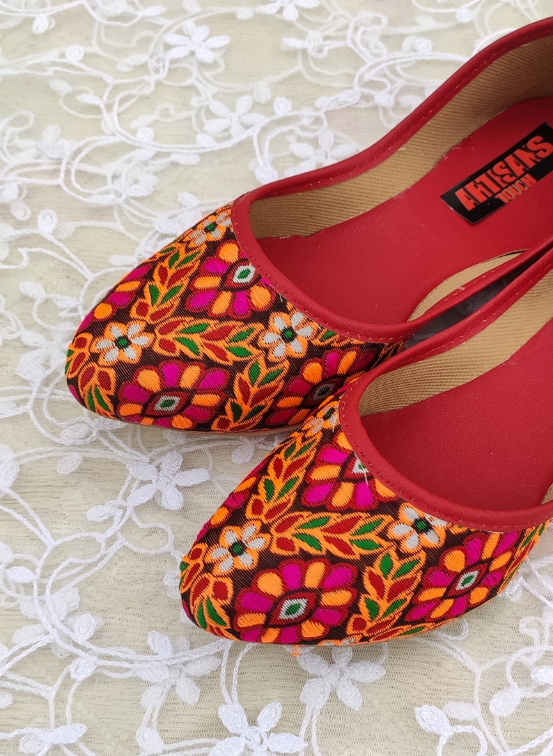 May include: Pair of red flat shoes with a colorful floral pattern. The shoes have a pointed toe and are made of fabric. The brand name "Artisans Touch" is visible on the insole of one shoe.