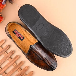 K&ouml;nnte beinhalten: Ein Paar braune Lederschuhe zum Hineinschl&uuml;pfen mit einem dekorativen Steppmuster. Die Schuhe haben eine schwarze Gummisohle und die Innensohle ist mit "Artisans Touch" beschriftet.