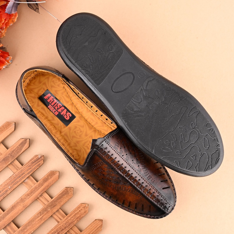 K&ouml;nnte beinhalten: Ein Paar braune Lederschuhe zum Hineinschl&uuml;pfen mit einem dekorativen Steppmuster. Die Schuhe haben eine schwarze Gummisohle und die Innensohle ist mit "Artisans Touch" beschriftet.
