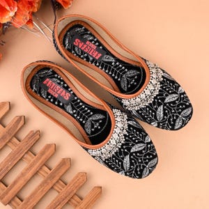 May include: A pair of black velvet flats with silver embroidery and embellishments. The shoes have a brown leather insole and the text "ARTISANS TOUCH" is printed on the insole.