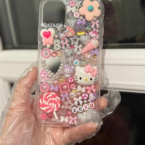 May include: Clear phone case adorned with pink and white decorations, including a Hello Kitty figure, bows, hearts, and a lollipop. The case also features the text "xoxo" and the Instagram handle "@hajra.s26."