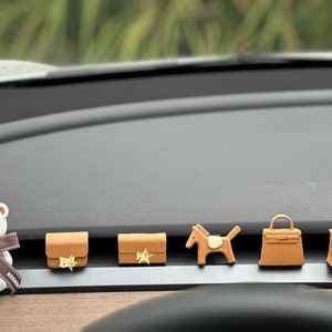 May include: A set of six brown car air freshener charms. The charms are shaped like handbags, a horse, and a teddy bear. The charms are all attached to a black dashboard.