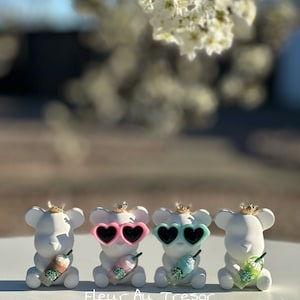 May include: Four white ceramic bear figurines wearing gold crowns and heart-shaped sunglasses. Each bear is holding a miniature iced coffee cup. The bears are arranged in a row on a white surface. The text "Fleur Au Tresor" is visible below the bears.