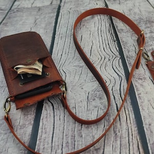 May include: A brown leather phone case with a strap. The case has a flap that opens to reveal a pocket for cash and cards. The strap is adjustable and can be worn over the shoulder.