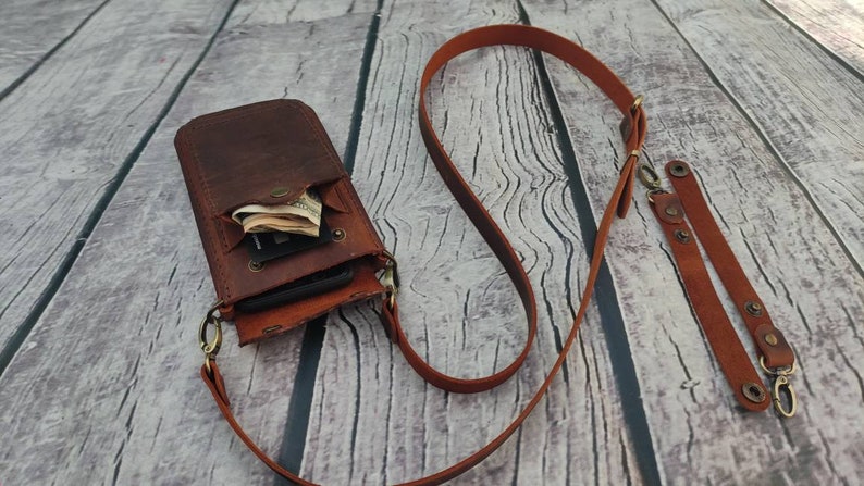 May include: A brown leather phone case with a strap. The case has a flap that opens to reveal a pocket for cash and cards. The strap is adjustable and can be worn over the shoulder.