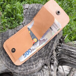 Puede incluir: Una cartera de cuero marrón con cierre a presión, abierta para revelar efectivo y una tarjeta de crédito. La cartera está sentada sobre un trozo de madera desgastada.
