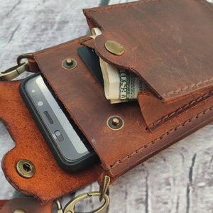 May include: A brown leather phone case with a flap that closes with a snap. The case has a strap that can be worn around the neck. The case is designed to hold a smartphone and has a small pocket for cash or cards.