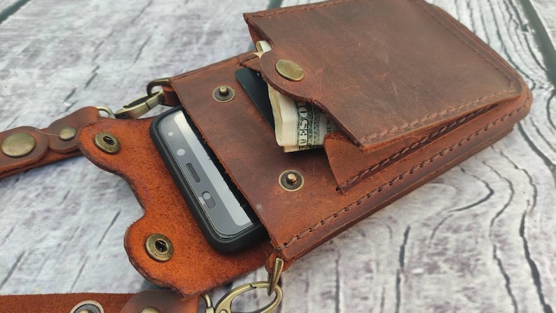 May include: A brown leather phone case with a flap that closes with a snap. The case has a strap that can be worn around the neck. The case is designed to hold a smartphone and has a small pocket for cash or cards.