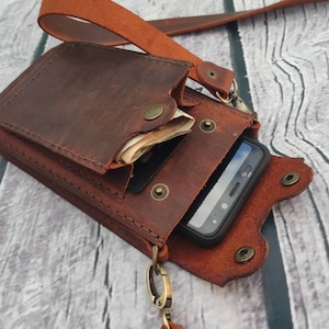 May include: A brown leather phone case with a flap that can be used to hold cash or cards. The case has a strap that can be worn over the shoulder.