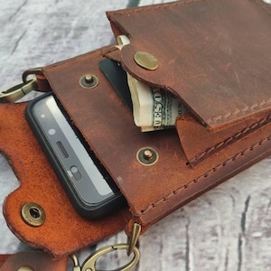 May include: A brown leather phone case with a flap that can be used to store cash or cards. The case has a strap that can be worn around the neck. The case is made of high-quality leather and is designed to protect your phone from scratches and bumps.