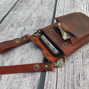 May include: A brown leather phone and wallet crossbody bag with a strap and a metal clasp. The bag has a pocket for cash and a separate pocket for a phone.