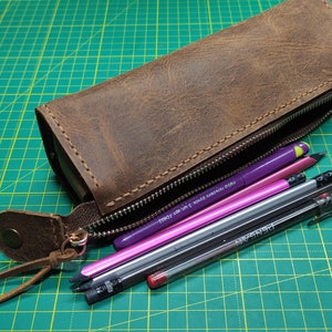 May include: A brown leather pencil case with a zipper closure and a leather strap. The case is filled with pens and pencils, including a purple pen with the text "TWSBI ECO - 0.5 mm - World Writing Instruments" on it.