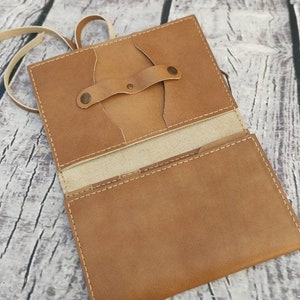 Puede incluir: Cartera de cuero marr&oacute;n claro abierta, con cierre de correa y bot&oacute;n. La cartera est&aacute; abierta, revelando bolsillos interiores y un forro de color claro. Costuras blancas detallan los bordes. Dise&ntilde;o sencillo y artesanal.