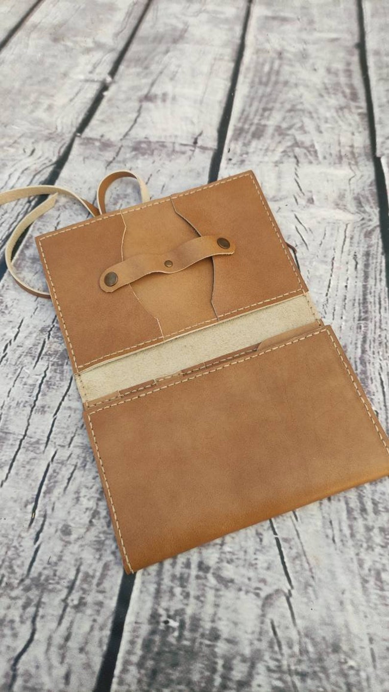 Puede incluir: Cartera de cuero marr&oacute;n claro abierta, con cierre de correa y bot&oacute;n. La cartera est&aacute; abierta, revelando bolsillos interiores y un forro de color claro. Costuras blancas detallan los bordes. Dise&ntilde;o sencillo y artesanal.