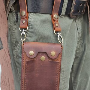 May include: A brown leather phone pouch with a flap and snap closure. The pouch is attached to a belt loop with two straps and metal clips.