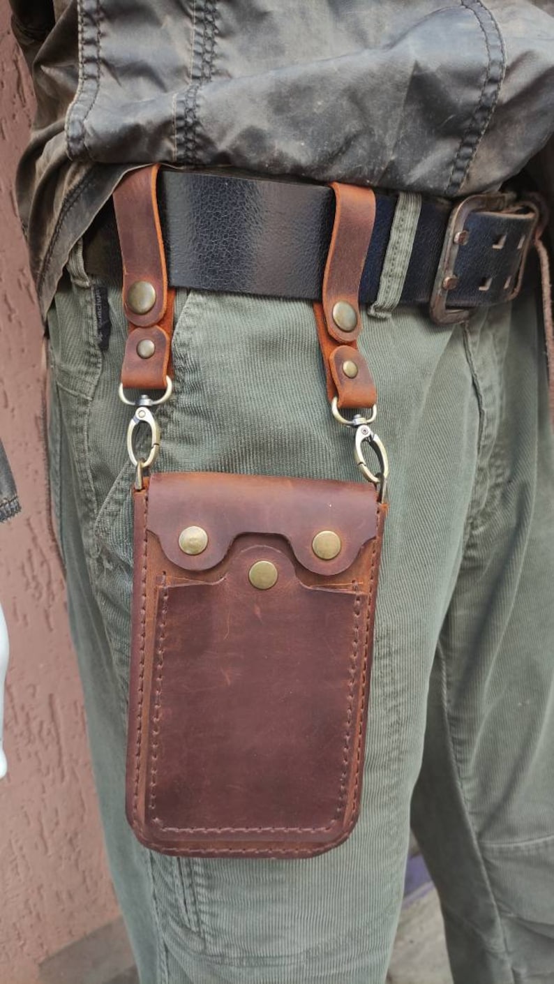 May include: A brown leather phone pouch with a flap and snap closure. The pouch is attached to a belt loop with two straps and metal clips.
