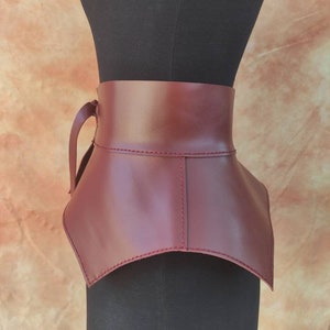 May include: A burgundy leather collar with a high neckline and a strap closure. The collar is displayed on a black mannequin, showcasing its shape and design. The leather appears smooth and the stitching is visible.