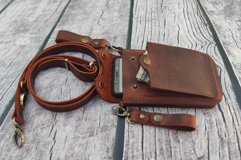 May include: A brown leather phone case with a strap and a pocket for cash. The case is designed to be worn around the neck or shoulder.
